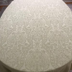 May include: An oval-shaped tablecloth in a light cream color featuring a repeating damask pattern. The fabric displays an intricate, woven design with symmetrical floral and scroll motifs that cover the entire surface of the textile.