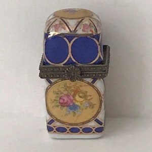 May include: A vintage porcelain sewing kit with a hinged lid. The kit features a blue cushion, a floral design, and gold accents. The oval-shaped floral design is centered on the front of the kit. The kit is a decorative and functional item.
