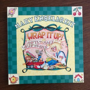 May include: A hardcover book titled "Wrap It Up! Gifts to Make, Wrap and Give" by Mary Engelbreit. The cover features a cartoon illustration of a person wrapping a gift, with a red wagon filled with gifts. The book has a green checkered border.