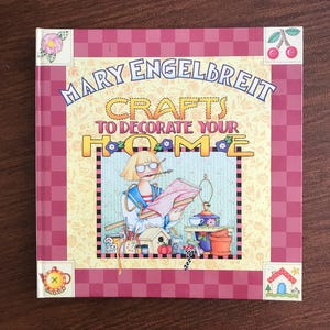May include: A square book cover with the title "Mary Engelbreit Crafts to Decorate Your Home." The cover has a pink and red checkered border. The illustration features a woman with glasses holding a drawing, surrounded by craft supplies.