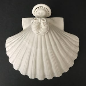 May include: A white ceramic angel figurine with a seashell-shaped dress. The angel wears a bonnet and has a bow at the chest. The figurine has a detailed, textured surface and is set against a dark background.
