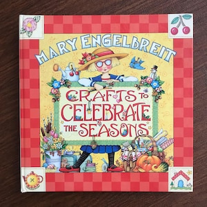 May include: A hardcover book titled "Crafts to Celebrate the Seasons" by Mary Engelbreit. The cover features a whimsical illustration of a girl in a hat, surrounded by seasonal crafts and a red checkered border.