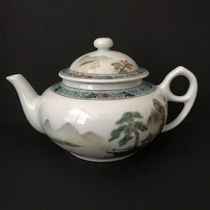 May include: A white porcelain teapot featuring a hand-painted landscape scene with green pine trees, mountains, and a small structure. The lid and shoulder are decorated with a teal and floral border. It has a curved spout and a looped handle.