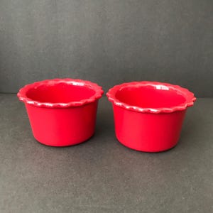 May include: Two red ceramic ramekins with scalloped edges. The ramekins are empty and sitting on a gray surface.