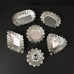 May include: A collection of six silver-colored metal baking molds in various shapes, including oval, round, diamond, and triangular forms. Each mold features a fluted, scalloped edge design with a reflective, metallic finish and a smooth center.