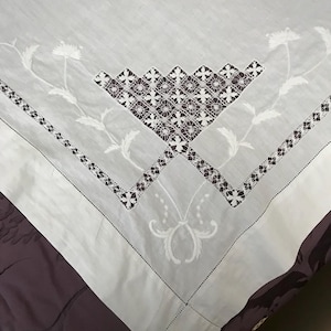 May include: White embroidered tablecloth with a geometric and floral design. The tablecloth features a central diamond pattern in brown and white, with floral accents and a decorative border. The fabric is draped over a dark purple surface.