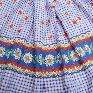 Op de afbeelding: Een kledingstuk van stof met een blauw-wit vichy-ruitpatroon en kleine rode hartjesmotieven. Een horizontale band over het onderste gedeelte toont een herhalend patroon van witte madeliefjes met gele harten op een blauwe achtergrond.