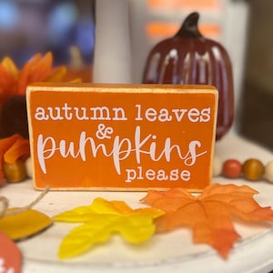 Puede incluir: Un bloque de madera naranja con las palabras "autumn leaves & pumpkins please" en escritura blanca. El bloque está rodeado de decoraciones otoñales, incluyendo hojas artificiales y una calabaza decorativa.