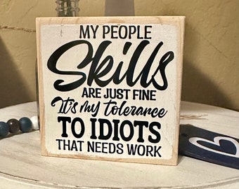 Op de afbeelding: Vierkant houten bord met de tekst "MY PEOPLE SKILLS ARE JUST FINE It's my tolerance TO IDIOTS THAT NEEDS WORK" in zwarte letters. Het bord heeft een verweerde, gebroken witte achtergrond en een natuurlijke houten lijst. Een decoratief item.