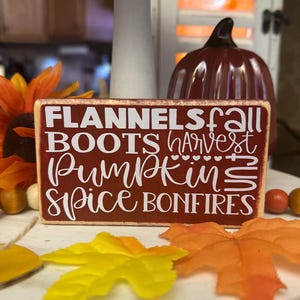 Könnte beinhalten: Ein rechteckiges Holzschild mit den Worten "FLANNELS, BOOTS, PUMPKIN, SPICE, BONFIRES" in weißer Schrift auf einem weinroten Hintergrund. Die Wörter "fall" und "harvest" sind ebenfalls enthalten. Künstliche Blätter und ein Kürbis im Hintergrund.