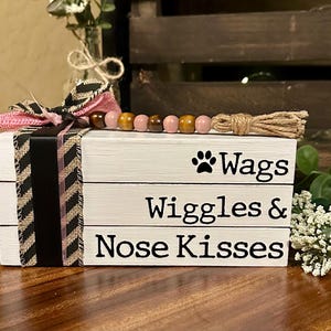 Puede incluir: Una caja de madera blanca con tres niveles, cada uno con un dicho diferente sobre perros. El nivel superior dice "Wags", el nivel intermedio dice "Wiggles &" y el nivel inferior dice "Nose Kisses". La caja está decorada con una cinta rosa y negra y una cadena de cuentas de madera.
