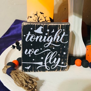 Puede incluir: Un letrero de madera cuadrado con un acabado negro desgastado y texto blanco que dice "tonight we fly". El diseño incluye un sombrero de bruja blanco, estrellas pequeñas, una escoba y la silueta de un murciélago. Los bordes muestran la veta.
