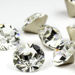 May include: Close-up of several clear, faceted gemstones with a brilliant cut. The gems are scattered, showcasing their reflective surfaces and the light they capture. The gemstones have a silver backing.