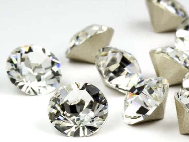 May include: Close-up of several clear, faceted gemstones with a brilliant cut. The gems are scattered, showcasing their reflective surfaces and the light they capture. The gemstones have a silver backing.
