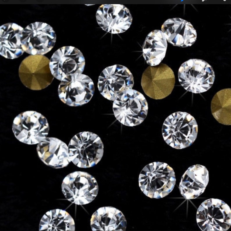 May include: Close-up of sparkling clear rhinestones and gold-colored metal studs scattered on a black surface. The rhinestones are round with multiple facets, reflecting light. The gold studs are also round and have a matte finish. Ideal for nail art or craft projects.