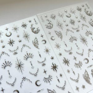 Könnte beinhalten: Zwei Bögen mit selbstklebenden Stickern, die eine Vielzahl von metallisch silbernen Designs aufweisen. Die Motive umfassen himmlische Elemente wie Sterne, Sonnen, Mondsicheln und stilisierte Flügel, die auf einer transparenten Folie angeordnet sind.