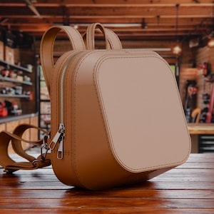 May include: A tan leather backpack with a rounded rectangular shape. The bag has a silver zipper, adjustable straps, and a top handle. The bag is sitting on a wooden surface.