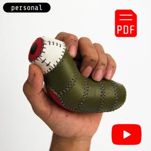 May include: A handcrafted, stitched leather object in olive green, white, and red, held in a hand. The object has a rounded shape with visible stitching. The word "personal" is above the object.
