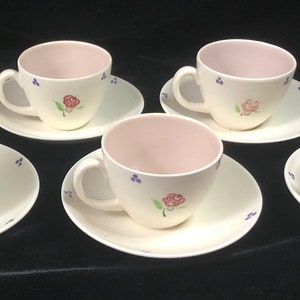 Puede incluir: Un juego de tazas y platillos de color crema con un diseño floral de rosas. El interior de las tazas es de color rosa claro. El juego incluye seis tazas y platillos, más una taza adicional. Las tazas y platillos están dispuestos sobre una superficie oscura.