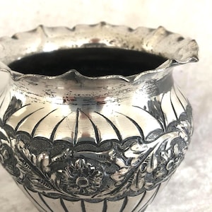 以下が含まれることがあります： フリル付きの縁が付いた、小さな銀色の丸い花瓶。花瓶の中央には詳細な花と葉の模様があり、底には垂直線があります。内部は暗いです。