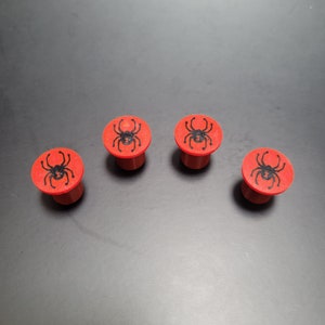May include: Four circular red plugs, each featuring a black spider graphic printed on the flat top surface. The plugs have a cylindrical base and a wider, flat circular head, arranged in a slight arc on a dark, neutral surface.