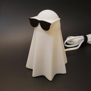 May include: A white ghost-shaped lamp wearing black sunglasses. The lamp has a rounded top and a draped body. A white cord is visible on the right. The lamp is on a black surface.