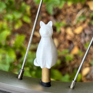 May include: A white, three-dimensional cat figurine attached to a brass-colored valve stem cap. The cat is seated in an upright position with pointed ears and a smooth surface texture. The object is positioned between two thin metal bicycle spokes.