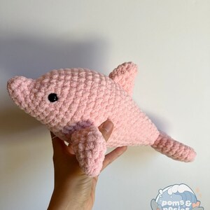 May include: A pink crocheted dolphin plush toy. The dolphin has a black eye and is made with a soft, fuzzy yarn.