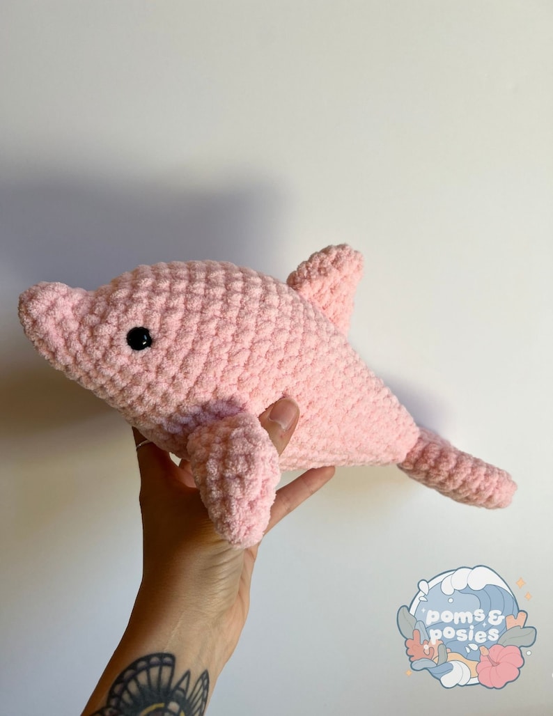 May include: A pink crocheted dolphin plush toy. The dolphin has a black eye and is made with a soft, fuzzy yarn.