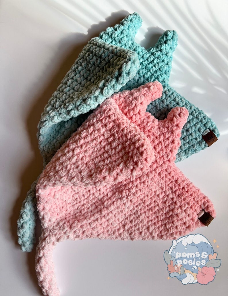 May include: Two crocheted hats, one in pink and one in blue, with a long tail. The hats are made with a fluffy yarn and have a textured surface.