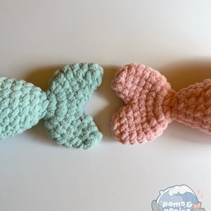May include: Two crocheted mermaid tails, one in light blue and one in pink, on a white background. The tails are made with a fluffy yarn and have a textured look.