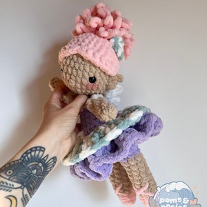 May include: A crocheted doll wearing a pink hat with a white and green striped bow, a purple dress with white and green stripes, and pink ballet shoes. The doll is holding a pink flower.