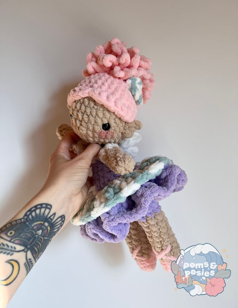 May include: A crocheted doll wearing a pink hat with a white and green striped bow, a purple dress with white and green stripes, and pink ballet shoes. The doll is holding a pink flower.