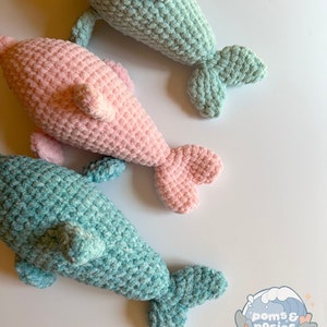 May include: Three crocheted dolphin plushies. One is pink, and two are light blue. The plushies are on a white background. The plushies have a textured, crocheted surface.