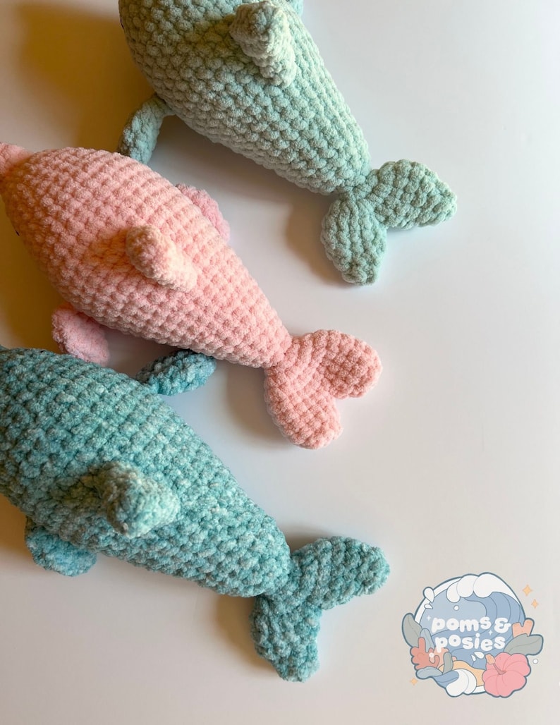 May include: Three crocheted dolphin plushies. One is pink, and two are light blue. The plushies are on a white background. The plushies have a textured, crocheted surface.