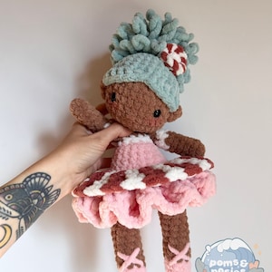 May include: A crocheted doll with brown skin, wearing a pink and white dress with a ruffled skirt, a blue hat with a candy cane accent, and pink ballet shoes. The doll has a curly blue yarn hairpiece.