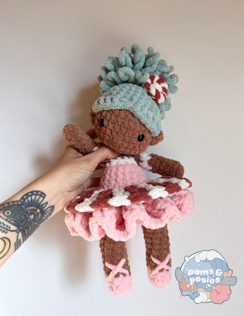 May include: A crocheted doll with brown skin, wearing a pink and white dress with a ruffled skirt, a blue hat with a candy cane accent, and pink ballet shoes. The doll has a curly blue yarn hairpiece.