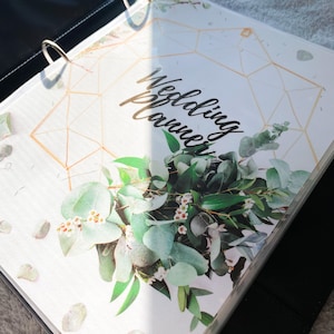 Puede incluir: Una carpeta blanca de planificación de bodas con un diseño geométrico dorado y una ilustración floral de hojas de eucalipto y flores blancas. El texto "Wedding Planner" está escrito en cursiva negra.