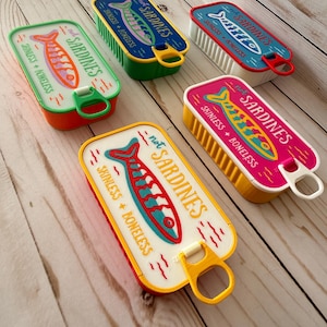 May include: Five rectangular containers shaped like sardine tins in various colors including green, blue, red, pink, and yellow. Each lid features a graphic of a fish and the text "not SARDINES" and "SKINLESS + BONELESS" with a pull-tab opener.