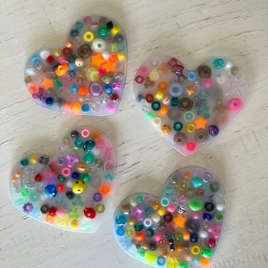 Puede incluir: Cuatro posavasos de resina en forma de corazón llenos de cuentas coloridas y pequeños adornos. Los posavasos presentan una variedad de formas y colores, incluyendo estrellas naranjas, caras sonrientes amarillas y varios tonos de azul, verde y rosa. Los posavasos están sobre una superficie blanca.