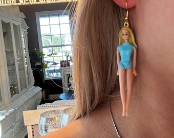 May include: A pair of gold dangle earrings featuring a miniature doll figure wearing a light blue swimsuit. The doll has blonde hair and a smiling face.