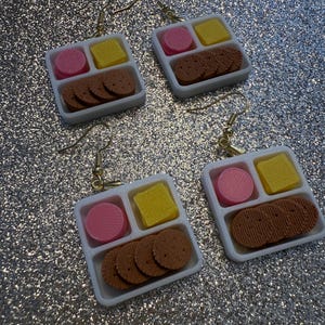 May include: Four pairs of novelty earrings. Each earring features a miniature white tray with a pink circle, yellow square, and brown biscuit-shaped pieces. The earrings have gold-colored hooks.