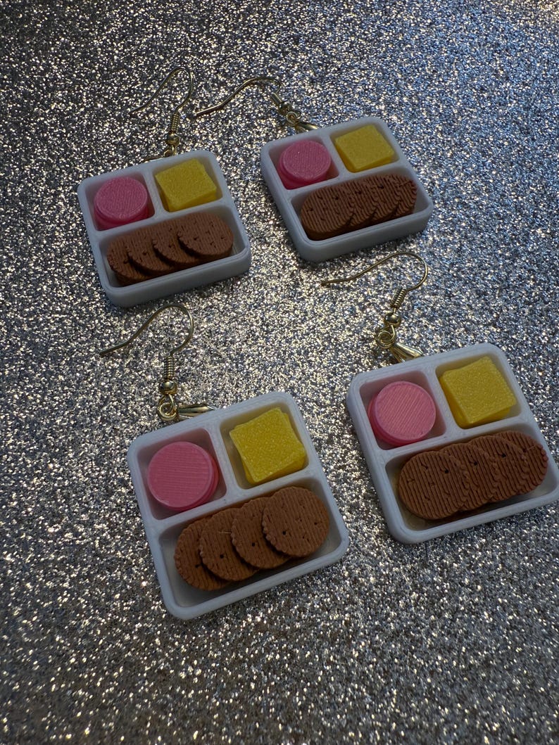 May include: Four pairs of novelty earrings. Each earring features a miniature white tray with a pink circle, yellow square, and brown biscuit-shaped pieces. The earrings have gold-colored hooks.