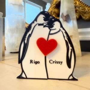 May include: A decorative penguin-themed item featuring two penguins embracing, with a red heart in the center. The penguins are black and white, with the names "Rigo" and "Crissy" below. The penguin on the left has a bow tie, and the penguin on the right has a necklace.