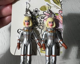 May include: A pair of silver astronaut doll earrings with brown boots and blonde hair. The earrings are on a white card with the text "Crissy's Creations".