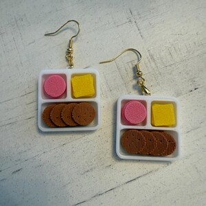 May include: A pair of novelty earrings shaped like miniature lunch trays. Each white tray has three compartments containing a pink circle, a yellow square, and several brown, round cookies. The earrings hang from gold-colored fish hook ear wires.