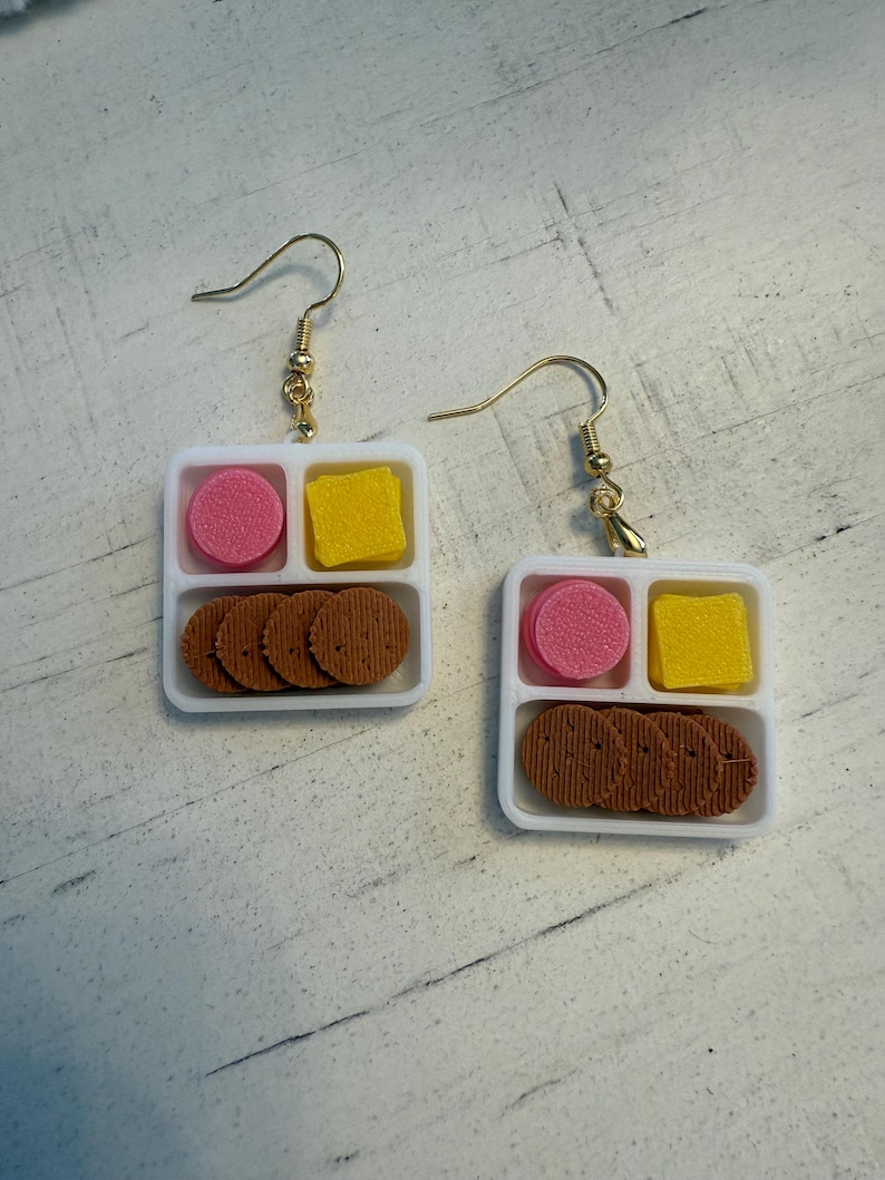 May include: A pair of novelty earrings shaped like miniature lunch trays. Each white tray has three compartments containing a pink circle, a yellow square, and several brown, round cookies. The earrings hang from gold-colored fish hook ear wires.
