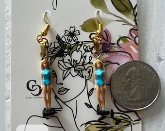 May include: Gold-toned dangle earrings featuring miniature doll figures in blue swimsuits. The earrings hang from gold-colored hooks. The card behind the earrings has the text "Crissy's Creations" and a drawing of a woman's face. A quarter is next to the earrings.