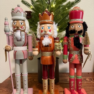 May include: Three wooden nutcrackers in a row, each with a unique design. The nutcrackers have painted faces, elaborate hats, and detailed uniforms in shades of pink, red, and orange. They stand on individual bases, with a Christmas tree in the background.