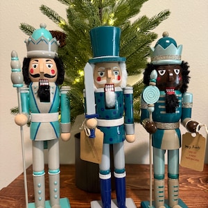 May include: Three decorative wooden nutcrackers in teal, gray, and blue. Each figure has a unique design, including a crown or top hat, and holds a staff or sword. They stand on painted bases, with a Christmas tree in the background.